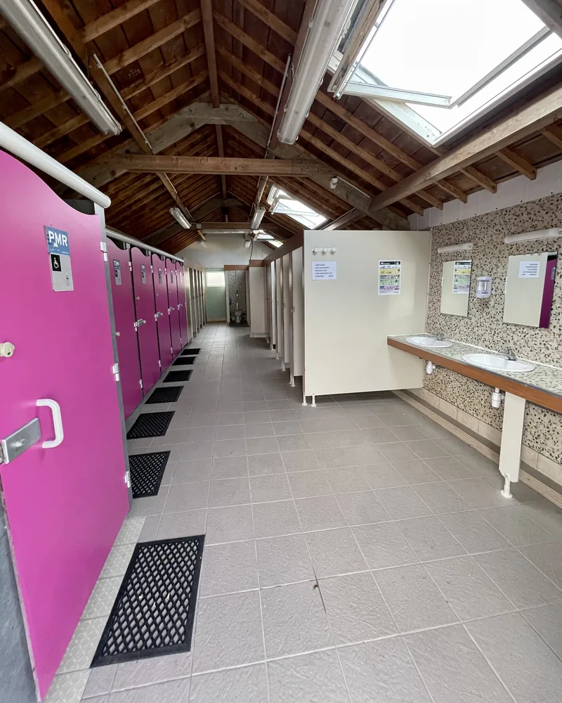 L’allée du bâtiment sanitaire : portes roses à gauche, dont une cabine PMR, lavabos à droite, charpente en bois et fenêtres de toit