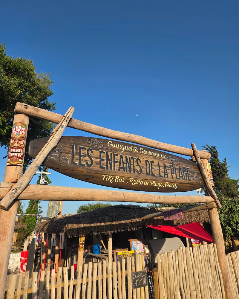 L’enseigne en bois de la guinguette, en forme de planche de surf : « Les Enfants de la Plage », entre deux totems tiki, sous un ciel bleu