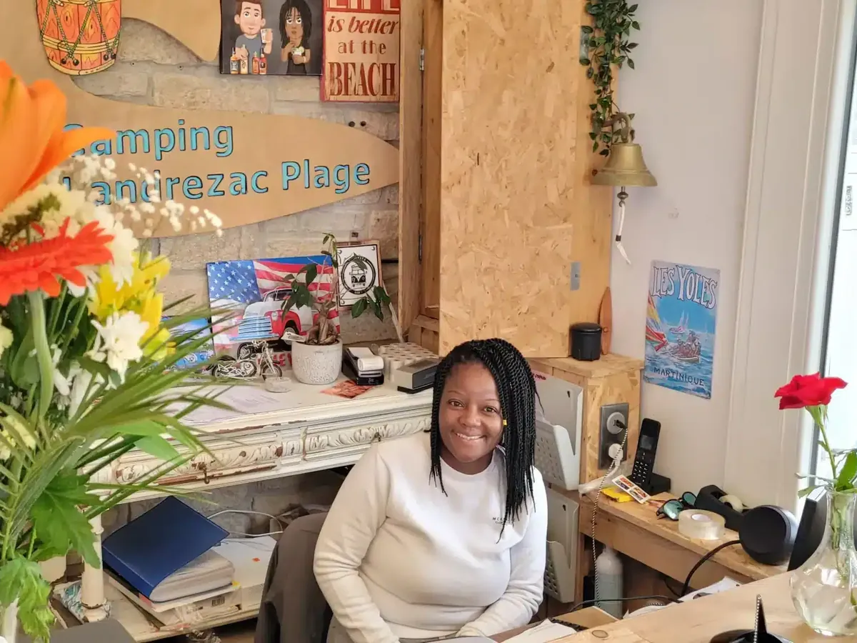 Mélye sourit, assise à la réception du camping ; devant elle, un bouquet de lys orange et, au mur, l’enseigne en bois « Camping Landrezac Plage »