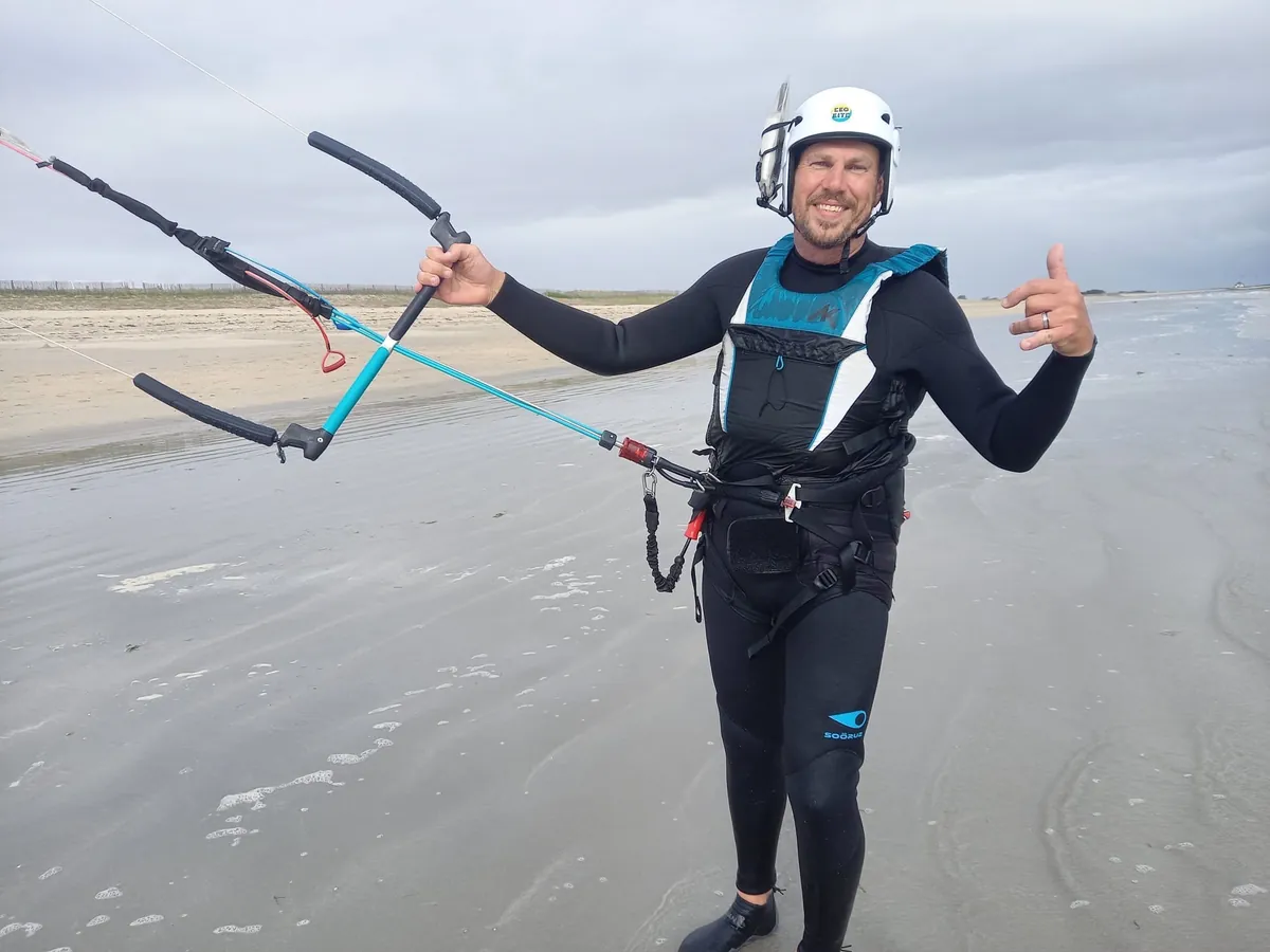 Fred sourit sur une plage à marée basse, en combinaison et casque de kitesurf, la barre de son aile à la main