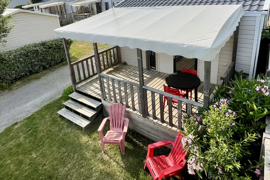 Le mobil-home Houat : terrasse en bois sous un auvent, deux fauteuils rouges sur la pelouse et des lauriers-roses en fleurs