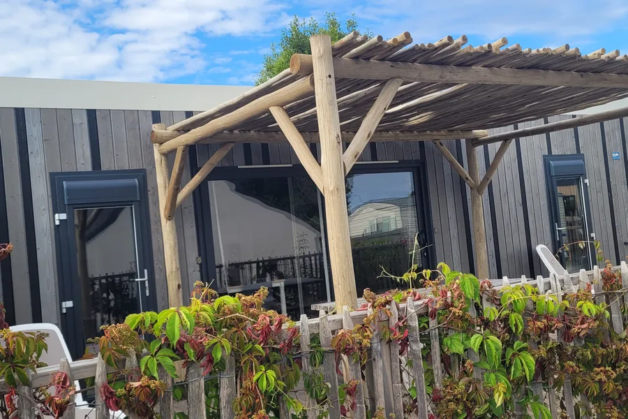 Le mobil-home Maldives, bardage gris, pergola en rondins et terrasse fermée par une ganivelle couverte de plantes grimpantes