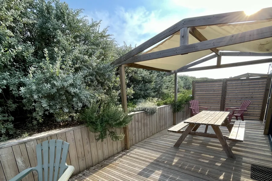 La terrasse en bois du mobil-home Moorea : une table de pique-nique sous une pergola à voile d’ombrage, des fauteuils et une haie d’arbustes