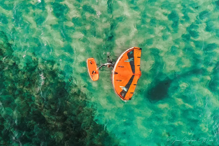 Vu du ciel : une personne en wing foil, aile orange à la main, sur une eau turquoise et transparente